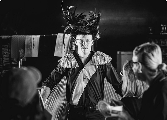A black and white photo of Jack Riewoldt dressed up for a television commercial. He wears a black satin blouse with a gold stripe, shoulder tassels and a black feather wig.