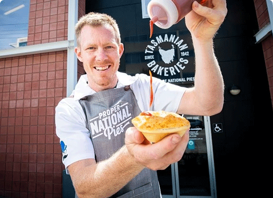 Jack Riewoldt posing with a pie as he squirts tomato sauce. He wears a blue National Pies branded apron and white collared shirt.