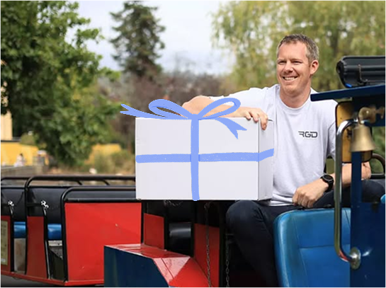 Jack Riewoldt smiling and sitting on a miniature train ride. He is holding a white package. He wears a plain white t-shirt.