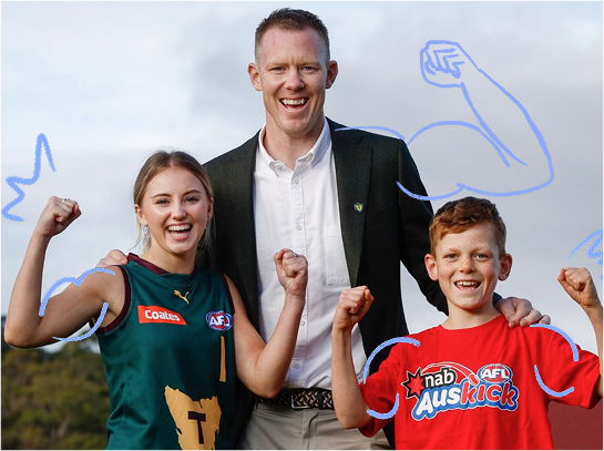 Jack Riewoldt with two young Tasmanian football fans—one in a Tasmania Devils guernsey and one in an AusKick t-shirt, both flexing and smiling